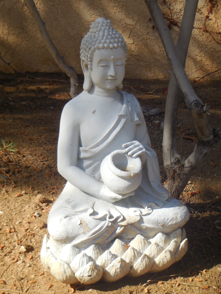 White statuette of a Thai monk, eyes closed, holding an empty vase, in contemplation. Set against bare tree limbs and brown dirt.