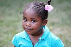 A young girl stares quizzically and beautifully into the camera; dressed in a bright turquoise shirt 