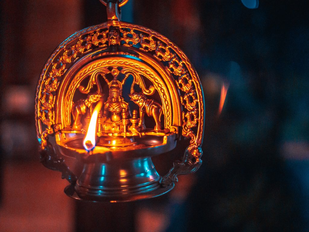 A single flame illuminating intricately carved candle holder, depicting Hindu god, praying figure