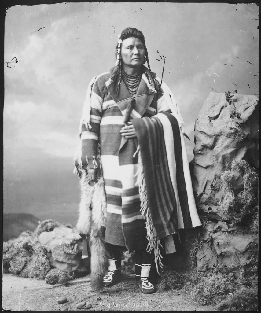 Chief Joseph of the Nez Perce, in ceremonial dress, hand over his solar plexus and serious, faraway aspect in his face.
