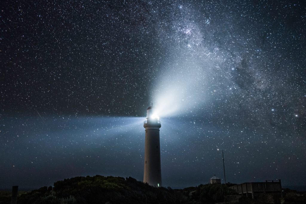 A lighthouse standing against stars in a dark sky, sending forth beams of light